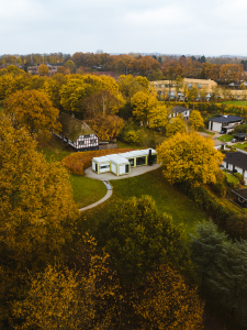 Foto taget fra drone, der viser et hvidt, modernistisk og rektangulært hus med fladt tag. Det er i en park med mange træer, hvis kroner har flotte, gule efterårsfarver. Bag det moderne hus ligger et gammeldags bindingsværkshus.