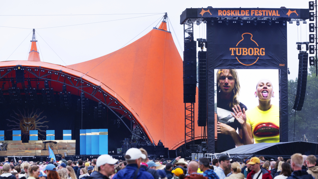 En menneskemængde står foran orange scene på Roskilde Festival. På en storskærm til højre for scenen ser man en ung mand og kvinde, der står foran en hvid baggrund. Kvinden rækker tunge.