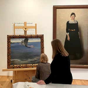 To kvinder henholdsvis står og sidder på hug foran maleriet Jomfru Blidelil på et staffeli. På væggen hænger det stor helfigurs portræt af kunstneren bag Jomfru Blidelil,