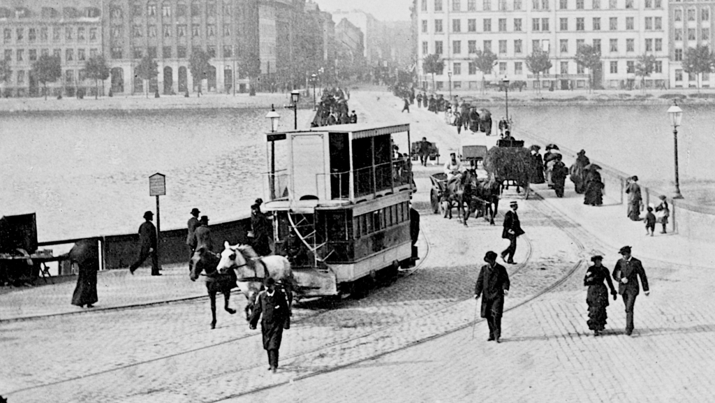 Sort hvidt foto af folk, der går over en bro over søerne i København. I forgrunden ses en hestetrukket sporvogn i to etager.