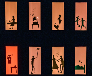 Sort flade med udskårne vinduer,med orangeagtigt lys i. I vinduerne ses sorte silhouetter af forskellige scenarier, fx en kvinde der sidder på en seng, en gammeldags grammofon med noder, der illustrerer at den spiller musik, to hunde der hopper rundt og leger og to børn der spiller bold.