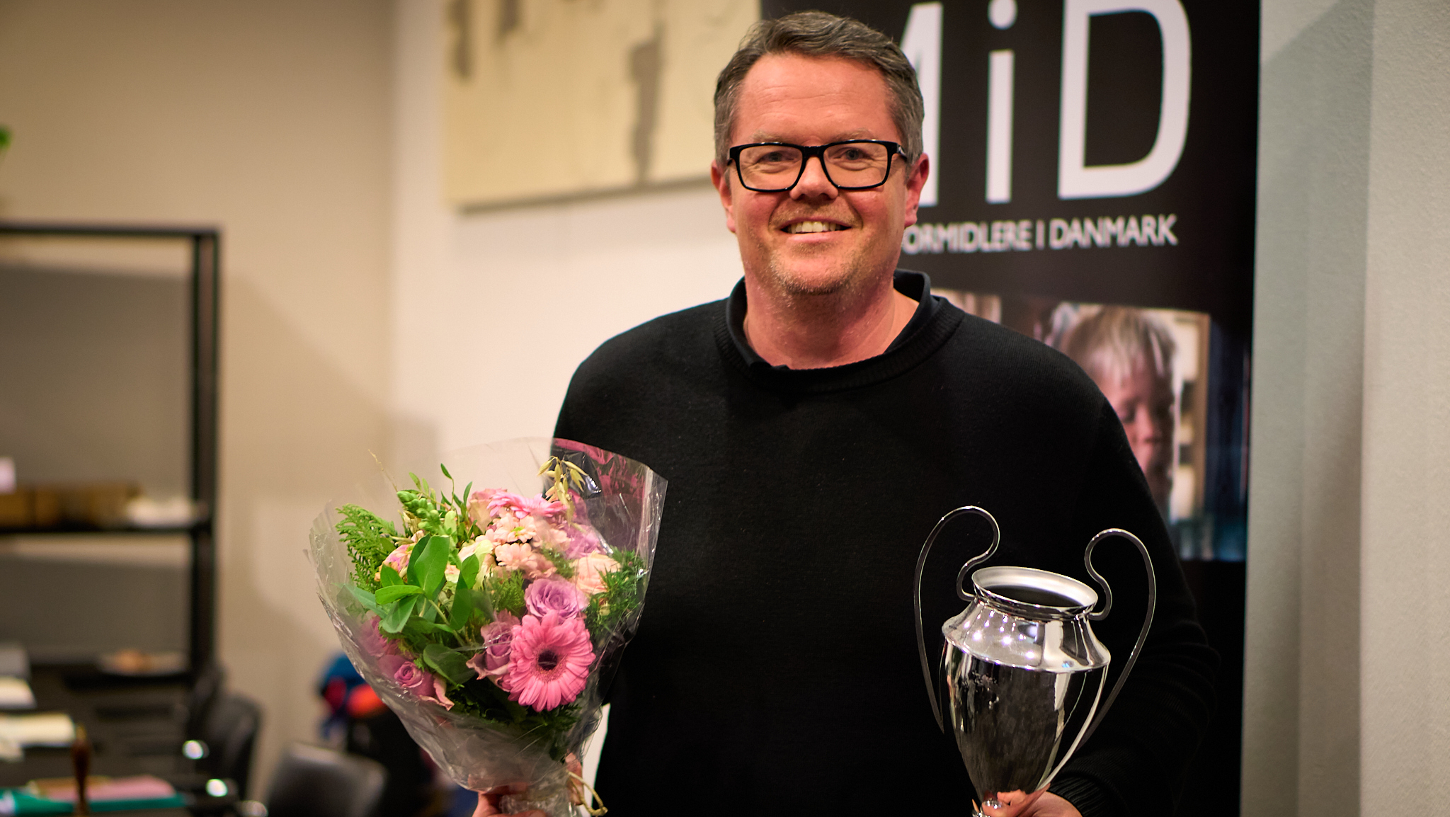 En mand med briller, kort hår og en sort trøje på smiller glad til kameraet. I den ene hånd holder han en buket blomster, i den anden holder han en sølvfarvet pokal.